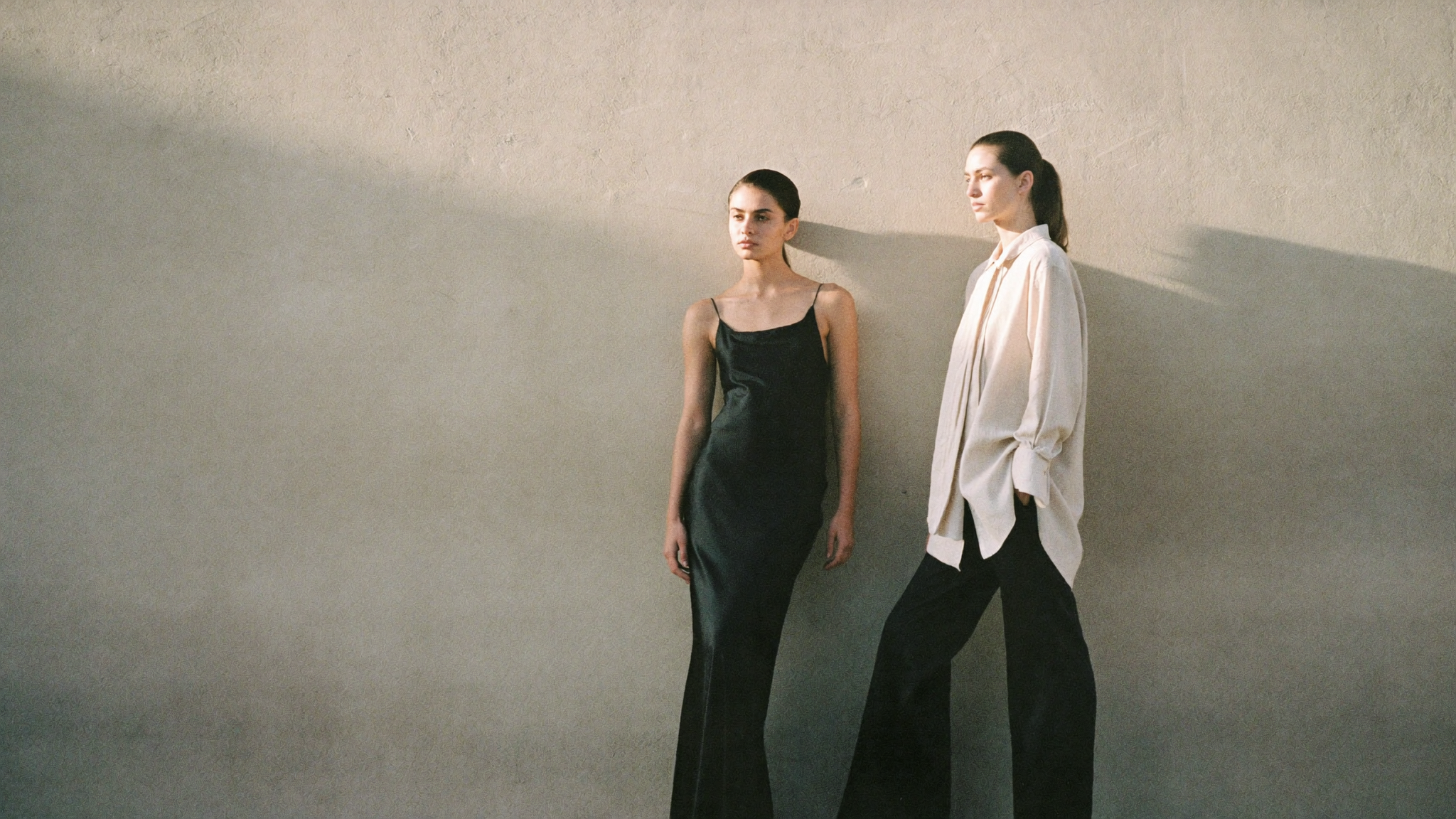 Novedades Hermosa — dos modelos ante muro de yeso beige, vestido de satén negro y camisa de lino ecru